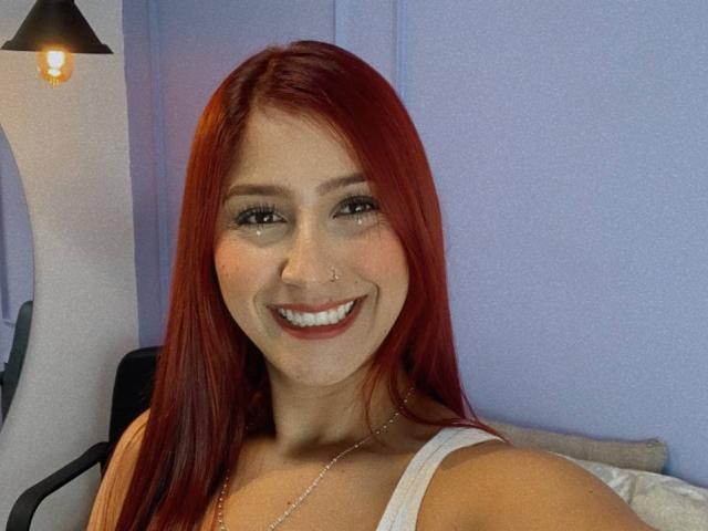 Lisa Harvie, com cabelo ruivo, sorrindo, vestindo uma camiseta branca, com um piercing no nariz e um colar, tem os seios levemente visíveis em uma selfie. O bumbum dela não está visível..