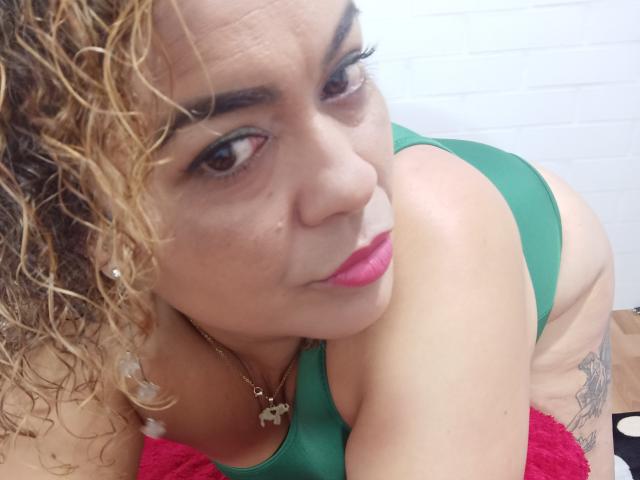 SolmaryHot with curly blonde hair, green one-piece, pink lipstick, looking up, showing her ass, tattoos on her left thigh, necklace with gold pendant, and pink towel.
