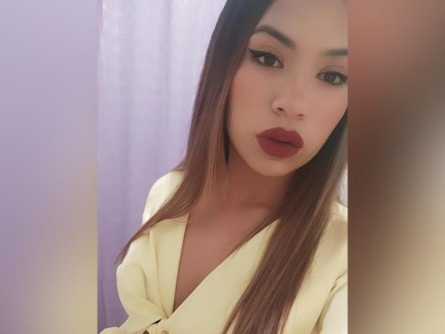ChloeDeluxee, avec de longs cheveux bruns, porte une chemise jaune à col en V, a du rouge à lèvres et regarde l'appareil photo avec une expression sérieuse. Ses seins sont partiellement visibles..