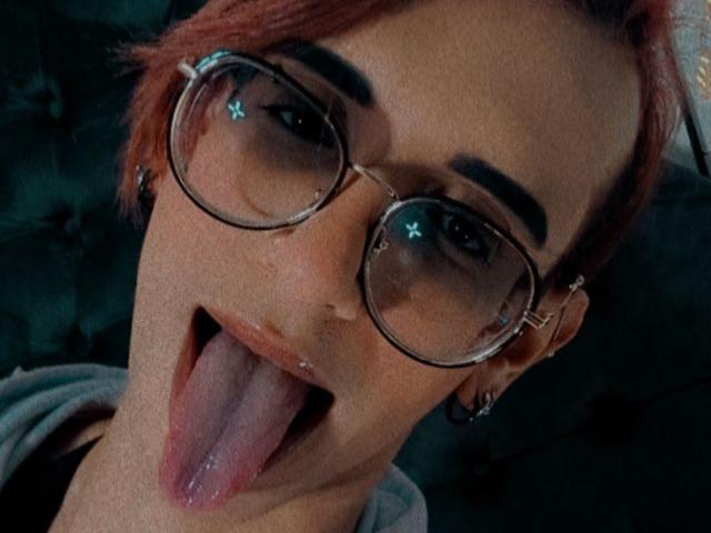 KrissyWyatt, com cabelo ruivo, usando óculos redondos, com a língua de fora, mostrando um lábio com piercing e piercings nas sobrancelhas. Fundo verde escuro..