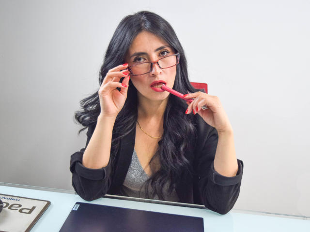JainaRogers, com longos cabelos pretos, usando óculos vermelhos e um casaco preto, chupando um lápis vermelho, segurando-o perto do nariz com unhas pintadas de vermelho. Seus seios estão visíveis..