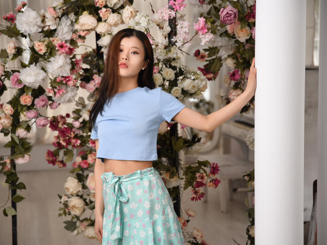 TinaKim with long brown hair, light blue crop top, and floral green skirt, standing against a white pillar, surrounded by pink and white roses. Her ass is slightly visible.