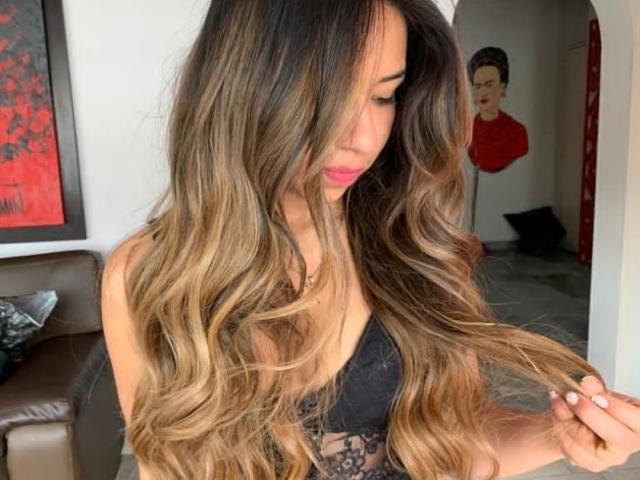 ChloeDeluxee, avec de longs cheveux bruns aux pointes blondes et ondulés, un soutien-gorge en dentelle noire, tenant une mèche de cheveux, rouge à lèvres rose, regardant vers le bas, dans une pièce moderne, en portrait..