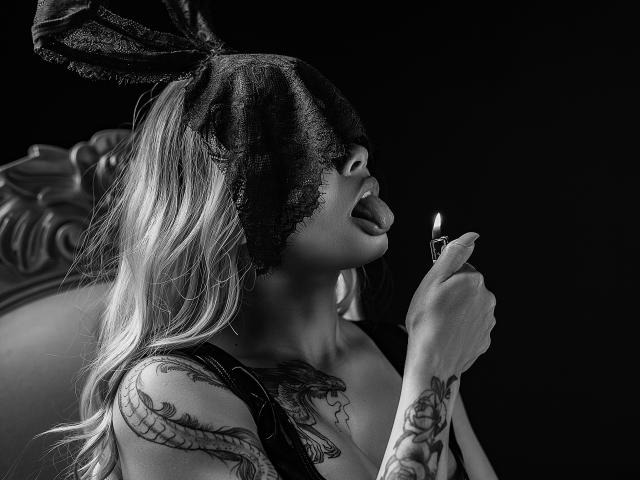 Black-and-white photo of DariaDawson with lace mask, tongue out, holding lit candle to her mouth. She's tattooed, with long blonde hair, and has visible breasts.