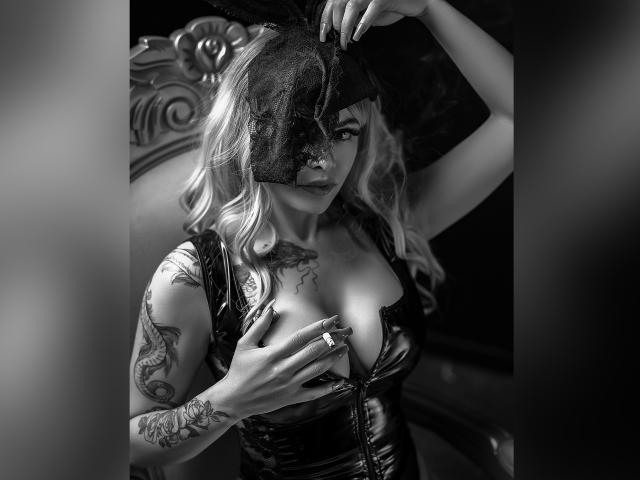 Black-and-white photo of DariaDawson with wavy blonde hair, wearing a sheer mask, leather corset, and finger ring, covering her tits with one hand, tattoos on right arm.