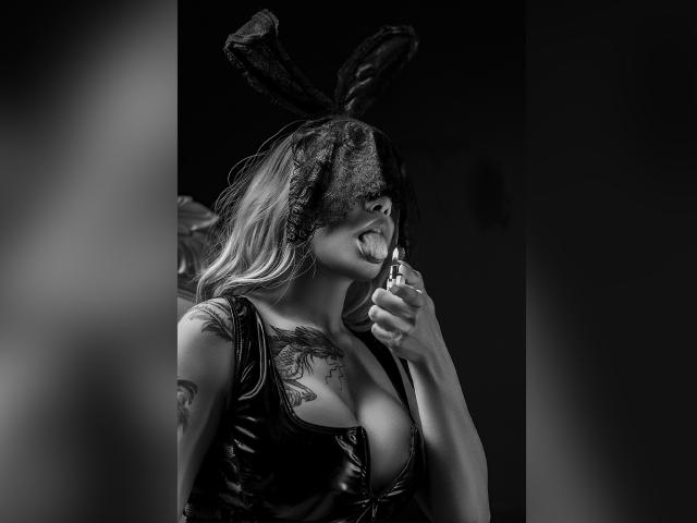 DariaDawson with blonde hair, black lace mask, and bunny ears, licking a lipstick applicator, wearing a shiny black latex top, showing large tits and a chest tattoo.
