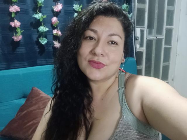 SakotoDika, con cabello negro rizado, lápiz labial rosa y una camiseta gris, aparece en un selfie mostrando sus pechos, sonriendo ligeramente. En el fondo, un sofá azul y flores rosas..