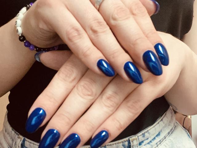 KarKutie's hands with shiny blue nail polish, fingers crossed, wearing a black top and purple bead bracelet, showing off her glossy, elongated nails.