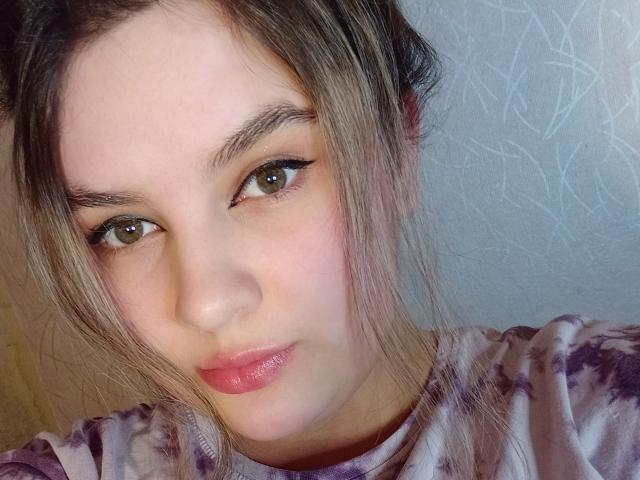 ConnyAlexandra's face with pink lips, brown eyes, and a slight smile. She has light brown hair in a messy bun, wearing a purple tie-dye shirt.