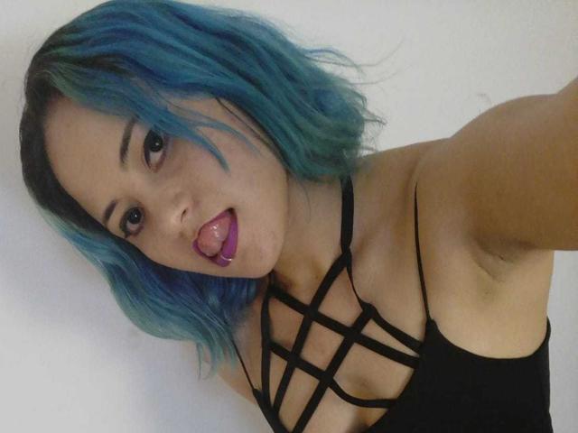 CassieJones with blue-green hair, black strappy top, tongue out, showing purple lipstick, and armpit visible, looking at camera. Boobs, tits, pussy not visible.