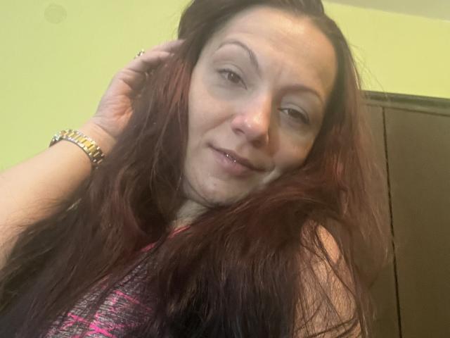 AamyBeatrice has long, dark red hair, a small nose ring, and a slight smile. She's wearing a pink and black plaid top, with a gold bracelet.