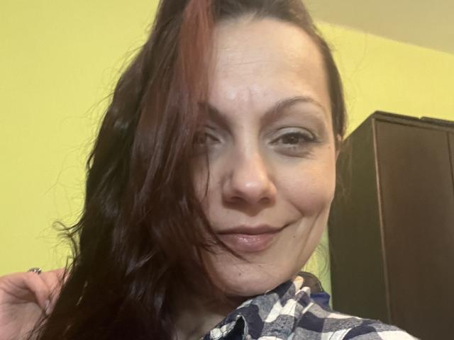 AamyBeatrice heeft een zelfvoldane glimlach, met golvend bruin haar dat gedeeltelijk haar gezicht bedekt. Ze draagt een geruite blouse en heeft subtiele make-up met opvallende eyeliner..
