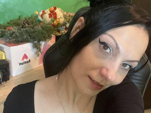 AamyBeatrice, met zwart haar, bleke huid en dikke zwarte eyeliner, zittend in een stoel, gekleed in een zwarte top, kijkend naar de camera met een lichte glimlach..