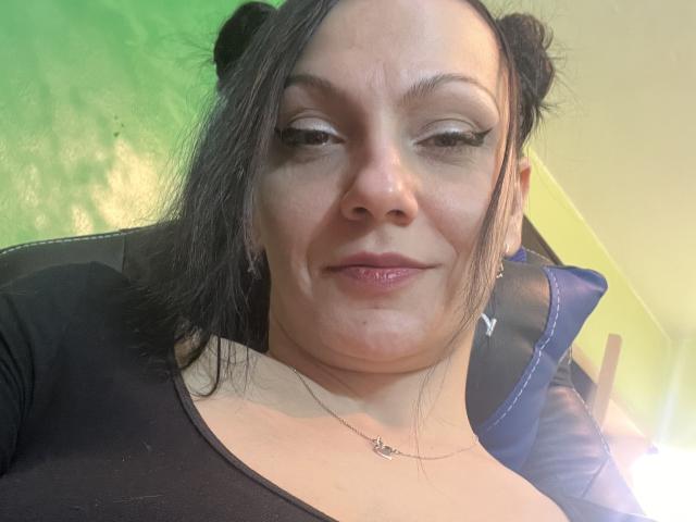 AamyBeatrice met zwarte, dubbele knotjes, glanzende oogmake-up en een subtiele grijns, gekleed in een zwarte top, zittend in een blauwe stoel, en met een kleine, glanzende mond..
