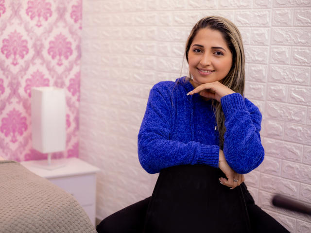 StefanyCaruso with blue sweater, black skirt, and long brown hair, smiling and resting her chin on her hand, showing off her cute ass. She has a ring on her right hand.