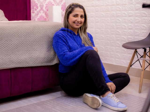 StefanyCaruso, zittend op de vloer, blauwe hoodie, zwarte broek, lichtblauwe sneakers, lachend, lang blond haar, witte bakstenen muur, roze en witte bed, houten stoel, ontspannen, schattig..