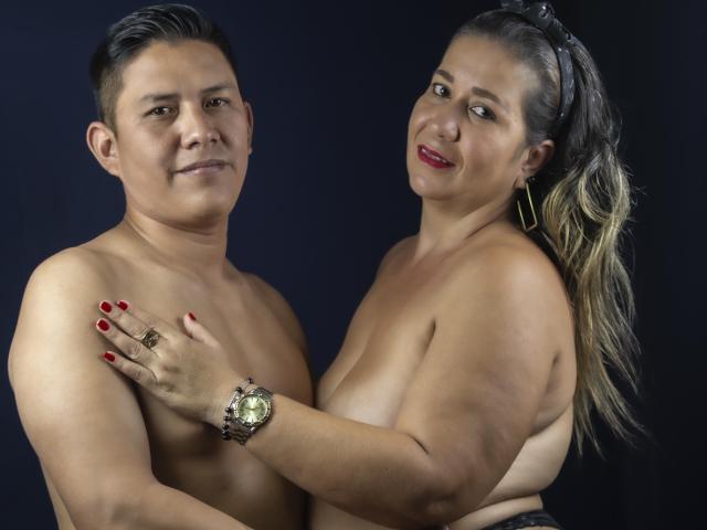 LeoGenesisCpl, een dik Aziatische man met kort zwart haar, zonder shirt, lachend, die een mollige Latina vrouw met lang blond haar, rode nagels en grote borsten vasthoudt, en die een horloge draagt..
