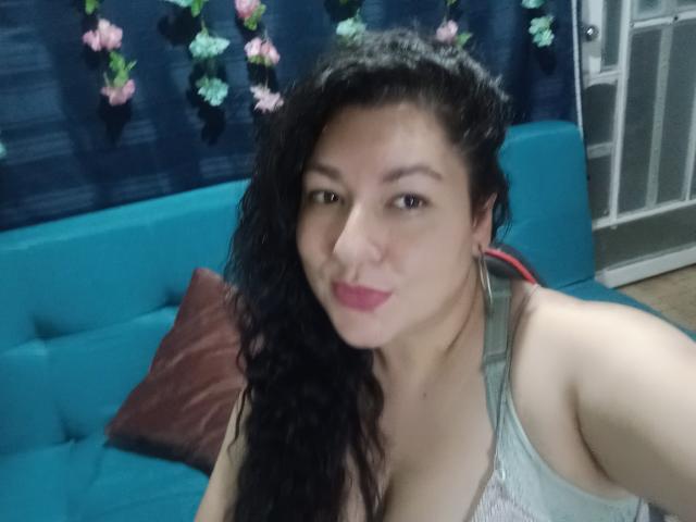 SakotoDika with wavy black hair, pink lipstick, and cleavage in a grey top, smiling at the camera, sitting on a blue couch with a brown pillow.