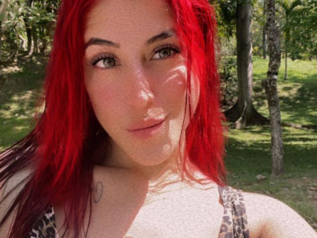 AlissaBrown with red hair, green eyes, light skin, small tattoo on right shoulder, wearing a leopard-print top, looking at camera in a sunny forest.