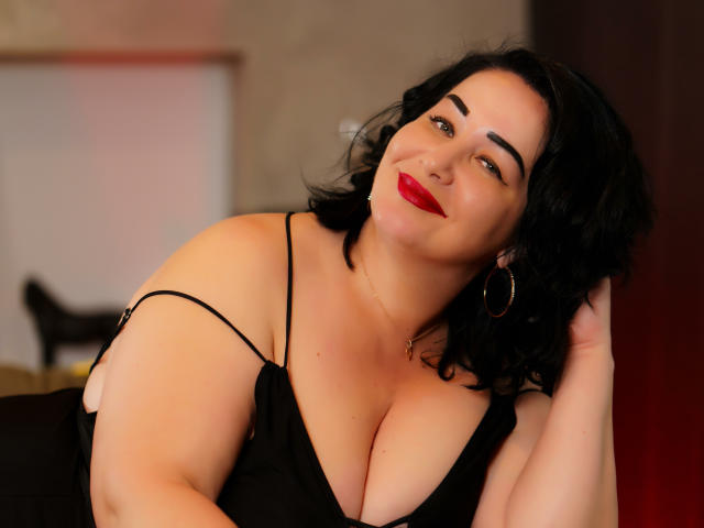 LexyRose with black wavy hair, red lipstick, and hoop earrings, smiling at camera, wearing a black dress with spaghetti straps, showcasing her large, round, and curvy boobs.