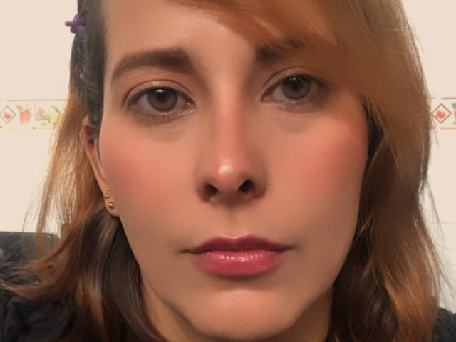 SaritaVelez's face, showing her brown eyes, pinkish lipstick, light brown skin, and shoulder-length brown hair with a purple hairclip. She's wearing small gold earrings.
