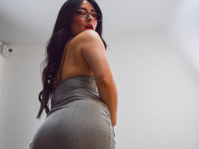 JainaRogers with long dark hair, glasses, and red lipstick, wearing a tight silver dress, showing her ass, looking back, low angle shot.