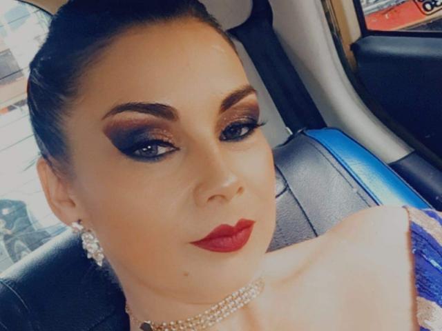 CruellaDeVil mit dunklem, rauchigem Augen-Make-up, rotem Lippenstift, einer Perlenkette, Diamant-Ohrringen und einem hochgesteckten Haarschnitt, sitzt in einem Auto und präsentiert ihr Hinterteil in einem blauen Kleid..