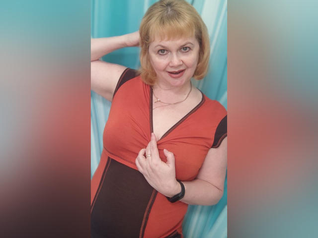 LilyHotLens con i capelli biondi corti, vestita con un abito rosso e nero, che tira giù l'abito per mostrare i suoi piccoli seni, sorridente, con un orologio da polso nero, in una foto..