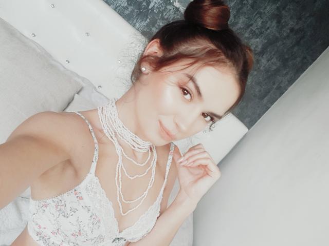 EmeraldFoxy with red hair in a bun, wearing a white lace bra and multiple pearl necklaces, smiling at the camera, her hand near her face, showing off her tits.