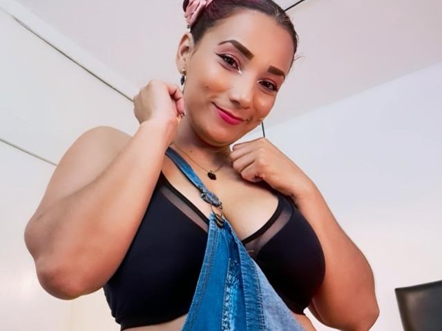 AmberDemons with pink hair, wearing a black bra, showing big tits, holding blue denim straps, smiling, pink lipstick, dark eyebrows, necklace, and pink ear gauges, in a selfie.