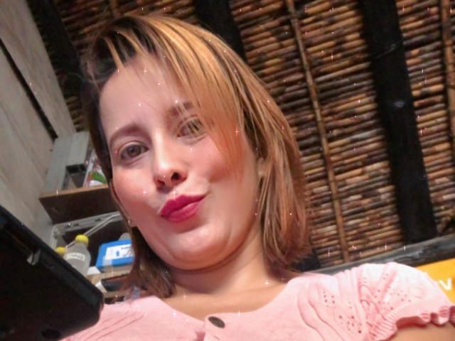 SaritaVelez with pink lipstick, puffy cheeks, short brown hair, wearing a pink shirt, looking into the camera with a slight pout, surrounded by subtle glitter effects.