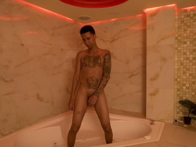 ElChicletv, desnudo, de pie en una bañera, tiene tatuajes en el pecho, los brazos y el muslo, con un pene circuncidado y flácido, y cabello corto y oscuro, en un baño con una iluminación cálida..
