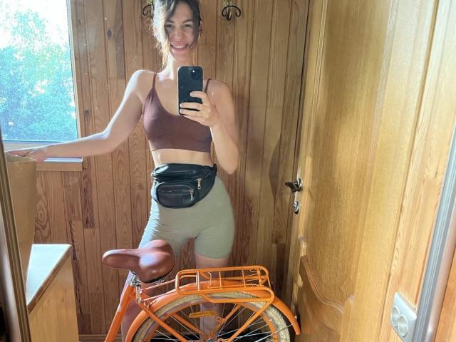 AurikaBriz, avec de petites seins, portant un soutien-gorge de sport marron, un short vert et une sacoche de taille noire, prend un selfie devant un miroir avec un vélo orange dans une pièce en bois..