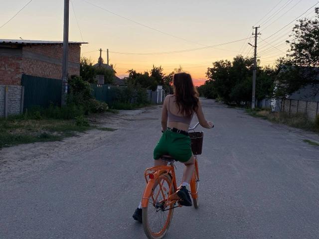 AurikaBriz with long brown hair rides an orange bike on a gravel road at sunset, wearing a white top and green shorts, with a brown basket on the bike.