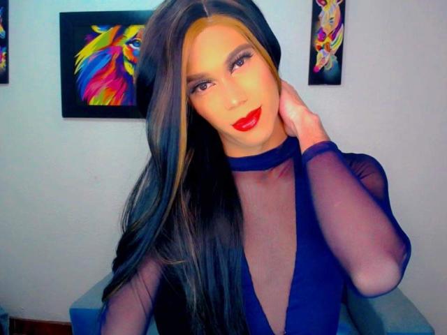 Mitchell Tay, con cabello largo, oscuro y con mechas rubias, vistiendo un vestido azul transparente, lápiz labial rojo, y mirando a la cámara, con la mano en el cuello, en una habitación muy iluminada..
