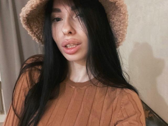 BellaMegan, con cabello negro largo, un sombrero rosa y una camisa marrón con textura, mostrando sus labios fruncidos y su pequeña nariz. Sus pechos están cubiertos por la camisa. Una sombra suave en la pared..