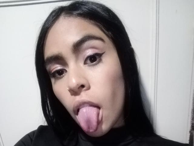 StellaBeckker has long black hair, brown eyes, and is sticking her pink tongue out. She's wearing black makeup with winged eyeliner. Her nose is pierced.