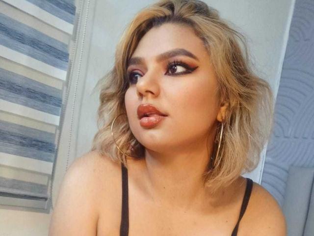 AliceeSecret hat blondes, welliges Haar, dunklen Eyeliner und glänzliche Lippen. Sie trägt große Reifenohrringe und ein schwarzes Oberteil und blickt zur Seite mit einem schmollenden Gesichtsausdruck..