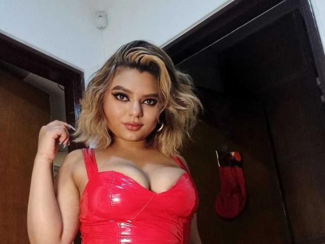 AliceeSecret mit blonden, gewellten Haaren trägt ein glänzendes, rotes Oberteil aus Vinyl, das tiefe Ausschnitte hat und ihre Brust betont. Sie hält ihre Haare und lächelt leicht in die Kamera. Ihre Brüste sind deutlich sichtbar..