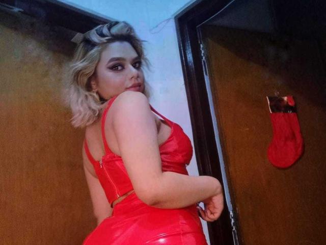 AliceeSecret with blonde hair, red leather lingerie, looking over her shoulder, showing her butt, in a dimly lit room with a red stocking on a wooden door.