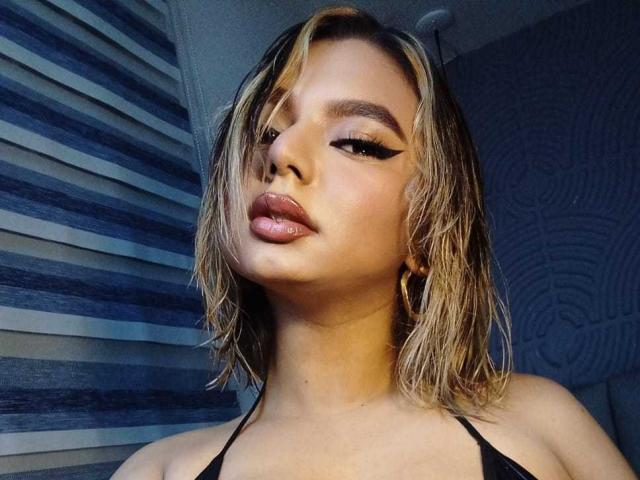 AliceeSecret con capelli biondi effetto bagnato, eyeliner scuro, labbra rosa lucide e orecchini a cerchio, indossa un top nero con spalline, guardando in basso con un'espressione leggermente corrucciata..