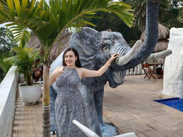 Maby Legrand de pie junto a una estatua detallada de un elefante gris, vestida con un vestido gris estampado, sonriendo, con una mano en la trompa de la estatua, y un árbol de palmera en el fondo..