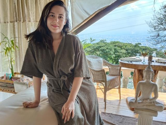 MabyLegrand, with dark brown hair and a cute smile, wearing a gray, textured bathrobe, leans on a beige cushion, next to a Buddha statue, by a sunny, wooden deck.