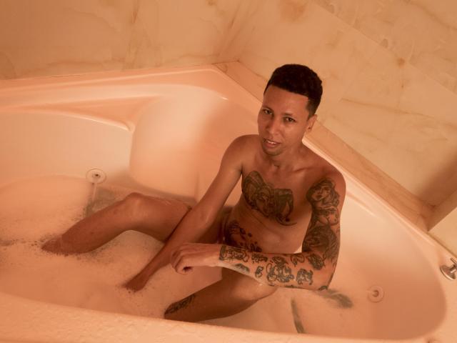 ElChicletv, naked with tattoos on chest and arm, sitting in a pink-tinted bathtub filled with bubbles, looking at the camera with a slight smile.