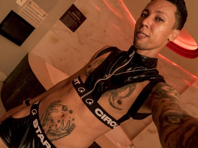 ElChicletv in black latex, showing tattoos, with a semi-nude chest and a visible stomach tattoo. Short, dark hair, smiling. Red light in the background.