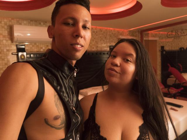 ElChicletv with short black hair and leather vest showing a tattoo, next to ElChicletv with long black hair and black lace bra, in a dimly lit room.