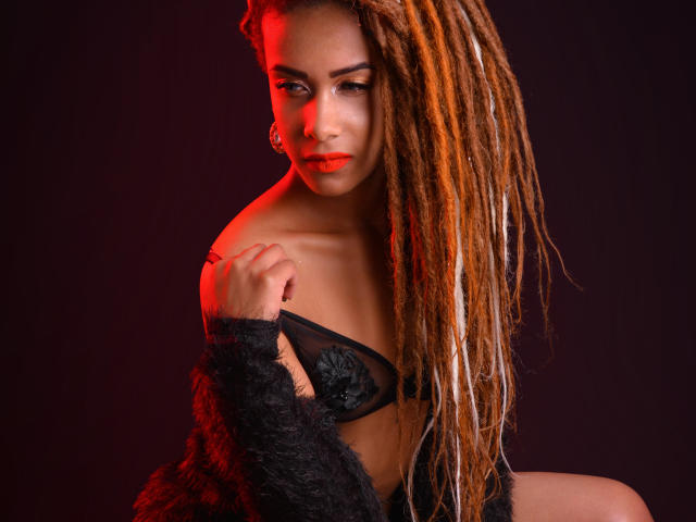 KendallQuinn with long, orange-brown dreadlocks, wearing a black, sheer bra with rose details, and a fuzzy black top, looking seductively at the camera, with red lighting.
