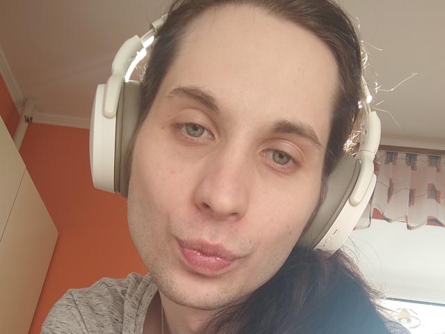 NikkiSandy, con ojos verde claro, usando auriculares blancos, con una ligera expresión de puchero, tiene un ligero bigote y cabello castaño recogido en una coleta, y lleva una camisa gris..