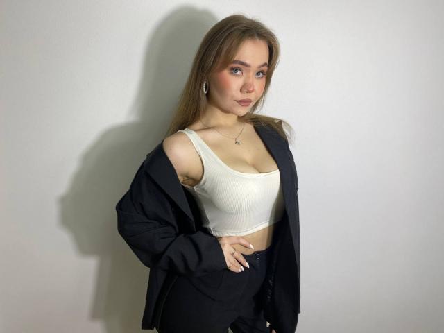 Stacy Yellig, avec de longs cheveux bruns, portant un débardeur blanc qui révèle ses seins, un pantalon noir et une veste noire, debout devant un mur blanc, regardant l'appareil photo..