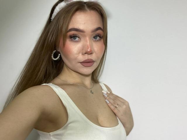 Stacy Yellig, con cabello castaño largo, vistiendo una camiseta blanca, mostrando senos pequeños, maquillaje rosado, una diadema blanca, un anillo y un arete de aro, sobre un fondo liso..
